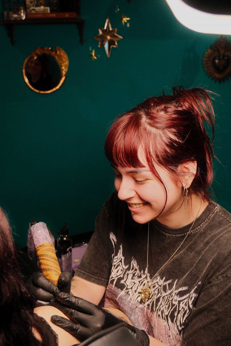 Inkzumi smiling as she tattoos in the studio, gloved hands holding the machine, with gilded mirrors on the deep green wall behind her